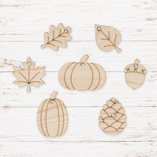 Seven unfinished light wood autumn-themed cutouts, including two pumpkins, a maple leaf, an oak leaf, a pine cone, and an acorn, arranged on a white distressed wood plank background.