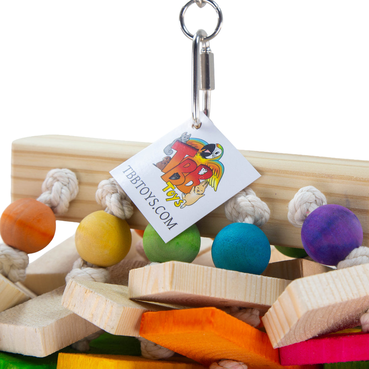 A close-up of a bird toy made of natural wood blocks, white rope, and colorful wooden beads in orange, yellow, green, blue, and purple. A small paper tag with the brand name 'TBBTOYS.