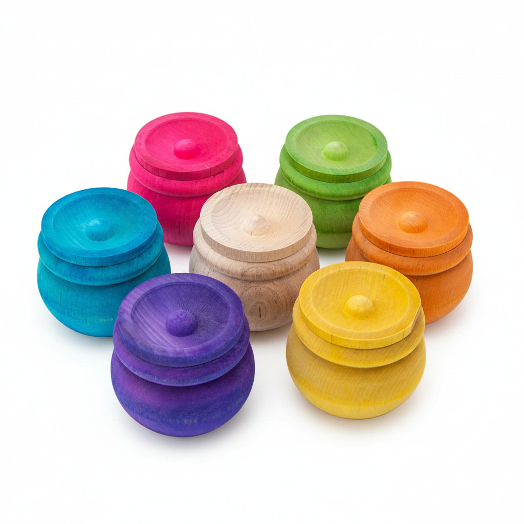 Seven small round wooden containers with lids, shown in assorted natural and  pink, green, blue, purple, orange, and yellow, arranged on a clean white background.
